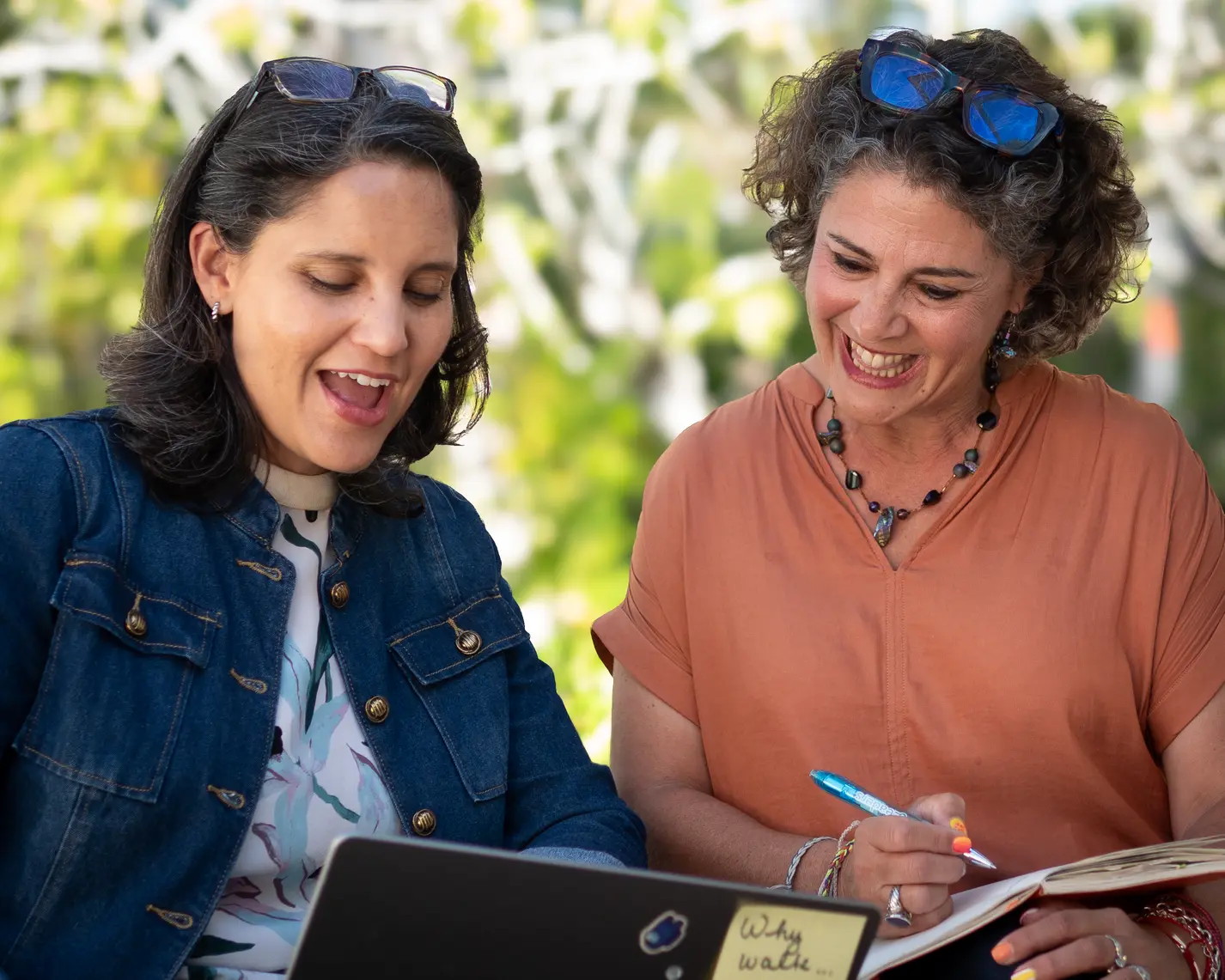 Two women collaborating outdoors over a laptop and notebook
